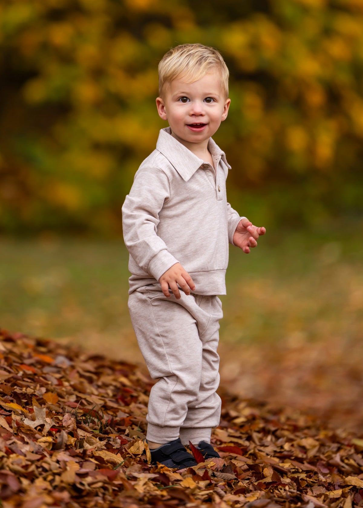 A young child with blonde hair stands on autumn leaves, wearing the Beckett and Bear Millie Brothers 2 PC Set with a collared neckline and dark shoes, set against a backdrop of blurred yellow and green foliage.