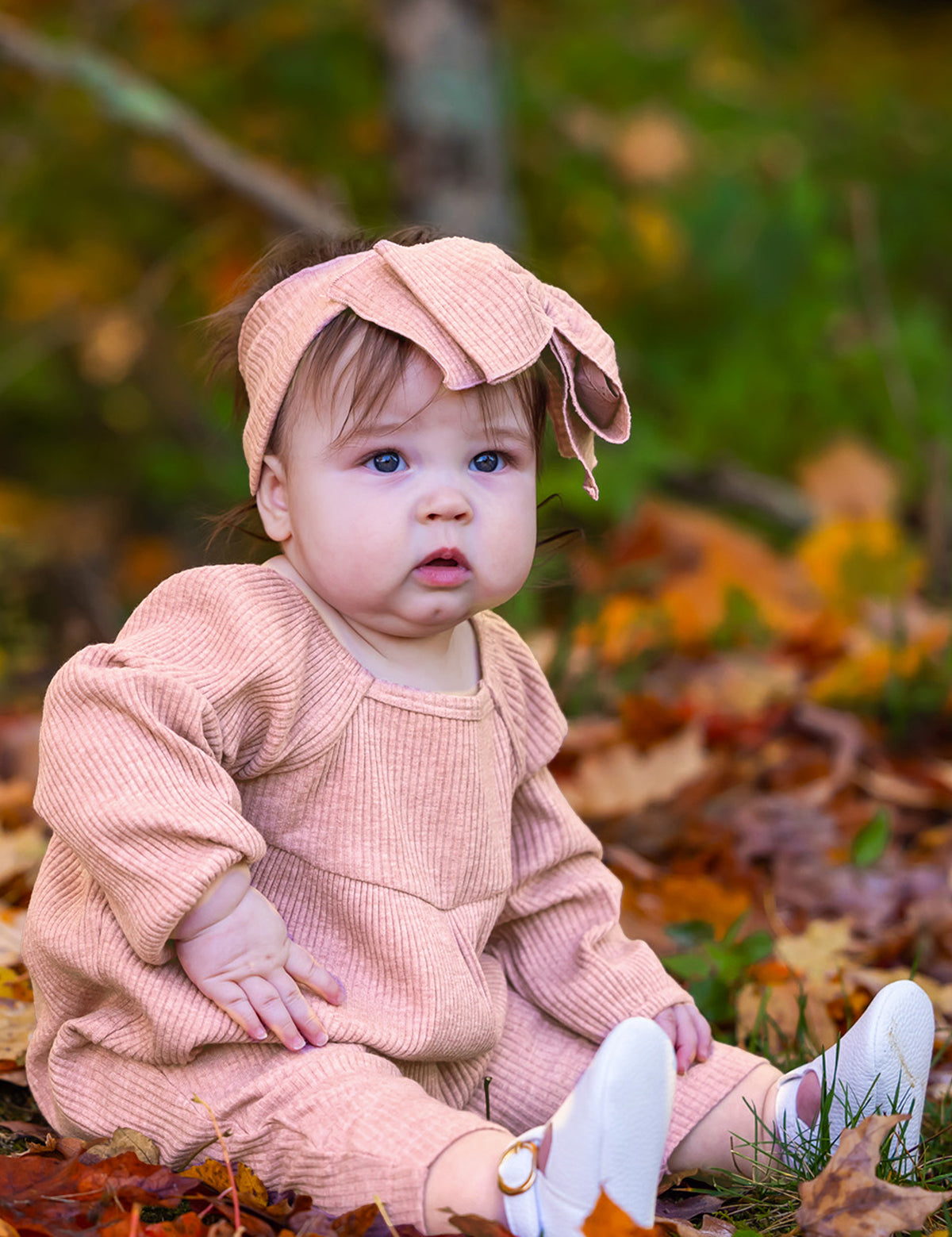 A baby in the Mabel + Honey Adore Baby Romper with a matching headband sits among autumn leaves, gazing curiously to the side.