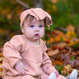 A baby in the Mabel + Honey Adore Baby Romper with a matching headband sits among autumn leaves, gazing curiously to the side.