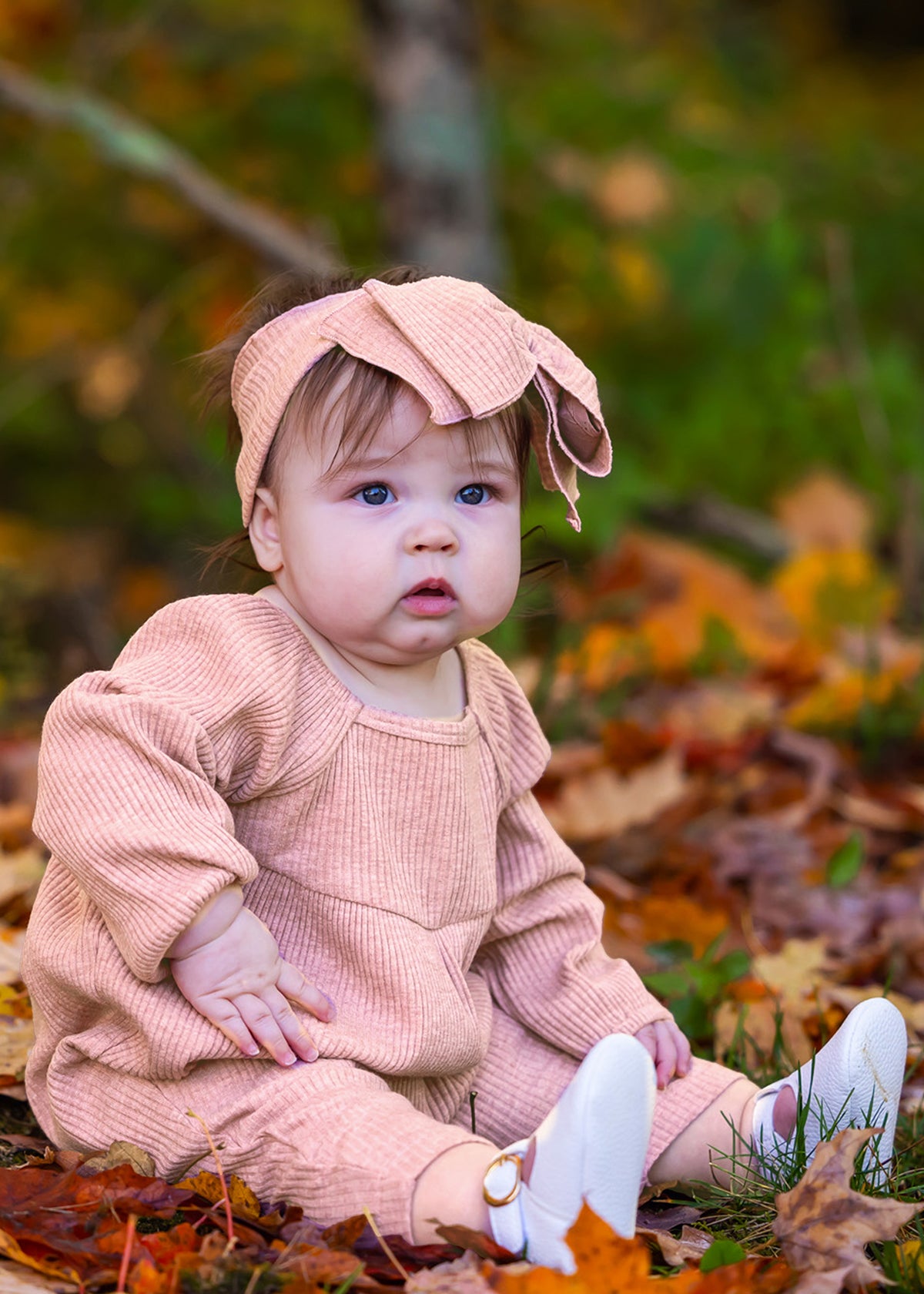 A baby in the Mabel + Honey Adore Baby Romper with a matching headband sits among autumn leaves, gazing curiously to the side.