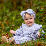 A smiling baby wears the Mabel and Honey Twinkle Twinkle Romper with ruffles and a matching large bow headband, sitting on grass surrounded by green foliage and autumn leaves.