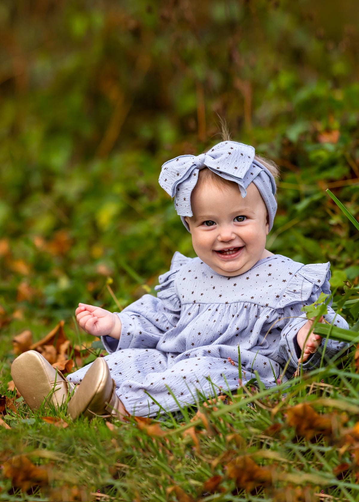 A smiling baby wears the Mabel and Honey Twinkle Twinkle Romper with ruffles and a matching large bow headband, sitting on grass surrounded by green foliage and autumn leaves.