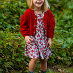 A young girl smiles outdoors in the Holly Dress by Mabel and Honey—a floral dress paired with a red cardigan, blue socks, and brown boots. She stands on green grass with leafy plants behind her.