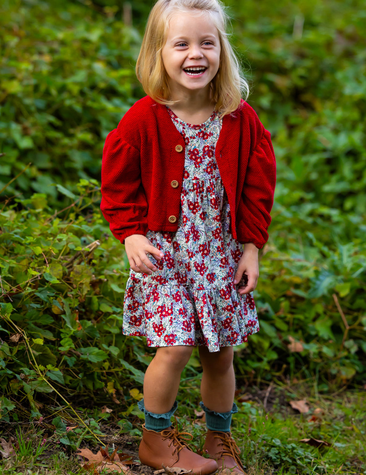 A young girl smiles outdoors in the Holly Dress by Mabel and Honey—a floral dress paired with a red cardigan, blue socks, and brown boots. She stands on green grass with leafy plants behind her.