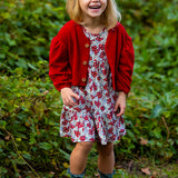 A young girl smiles outdoors in the Holly Dress by Mabel and Honey—a floral dress paired with a red cardigan, blue socks, and brown boots. She stands on green grass with leafy plants behind her.