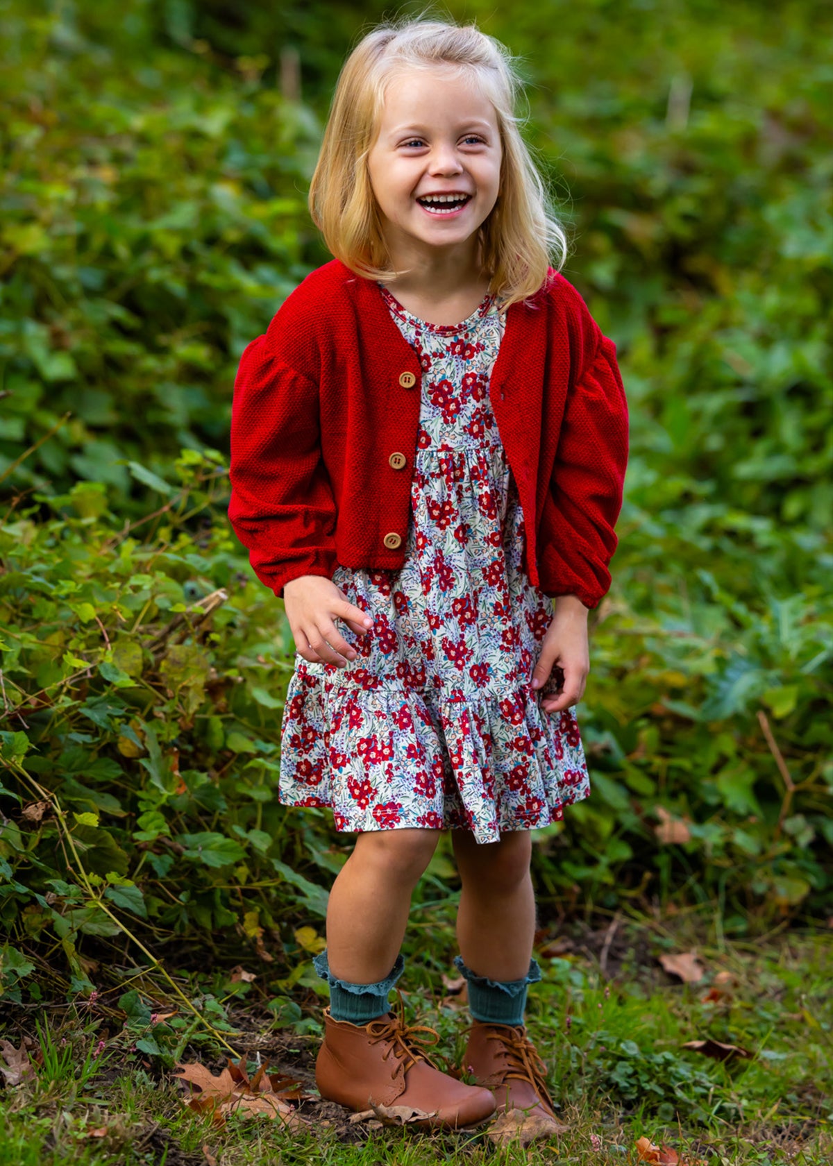 A young girl smiles outdoors in the Holly Dress by Mabel and Honey—a floral dress paired with a red cardigan, blue socks, and brown boots. She stands on green grass with leafy plants behind her.
