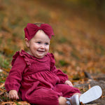 A smiling baby wears the Mabel and Honey Autumn Romper with a ruffled collar and matching headband, sitting on autumn leaves with trees softly blurred in the background.