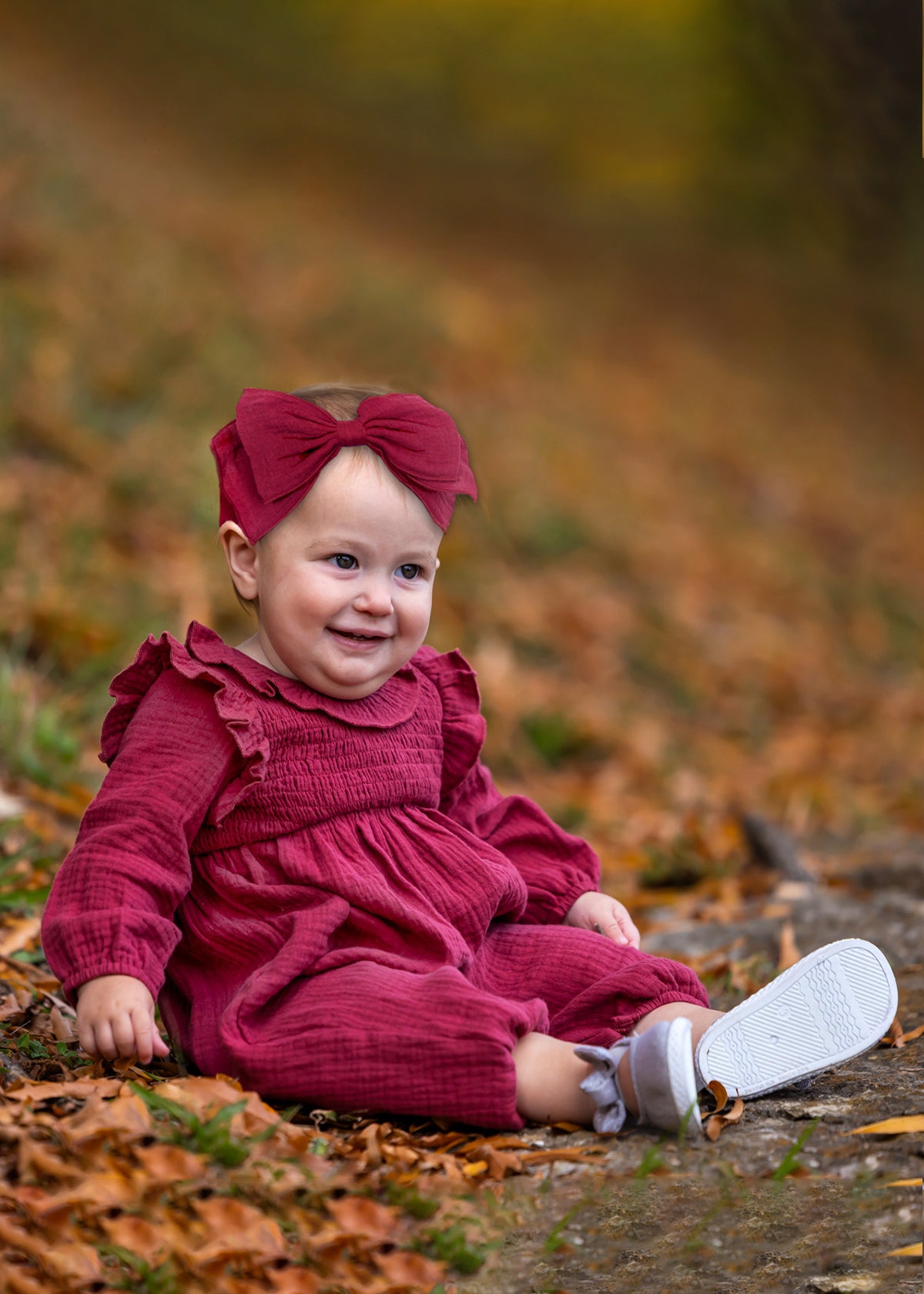 A smiling baby wears the Mabel and Honey Autumn Romper with a ruffled collar and matching headband, sitting on autumn leaves with trees softly blurred in the background.