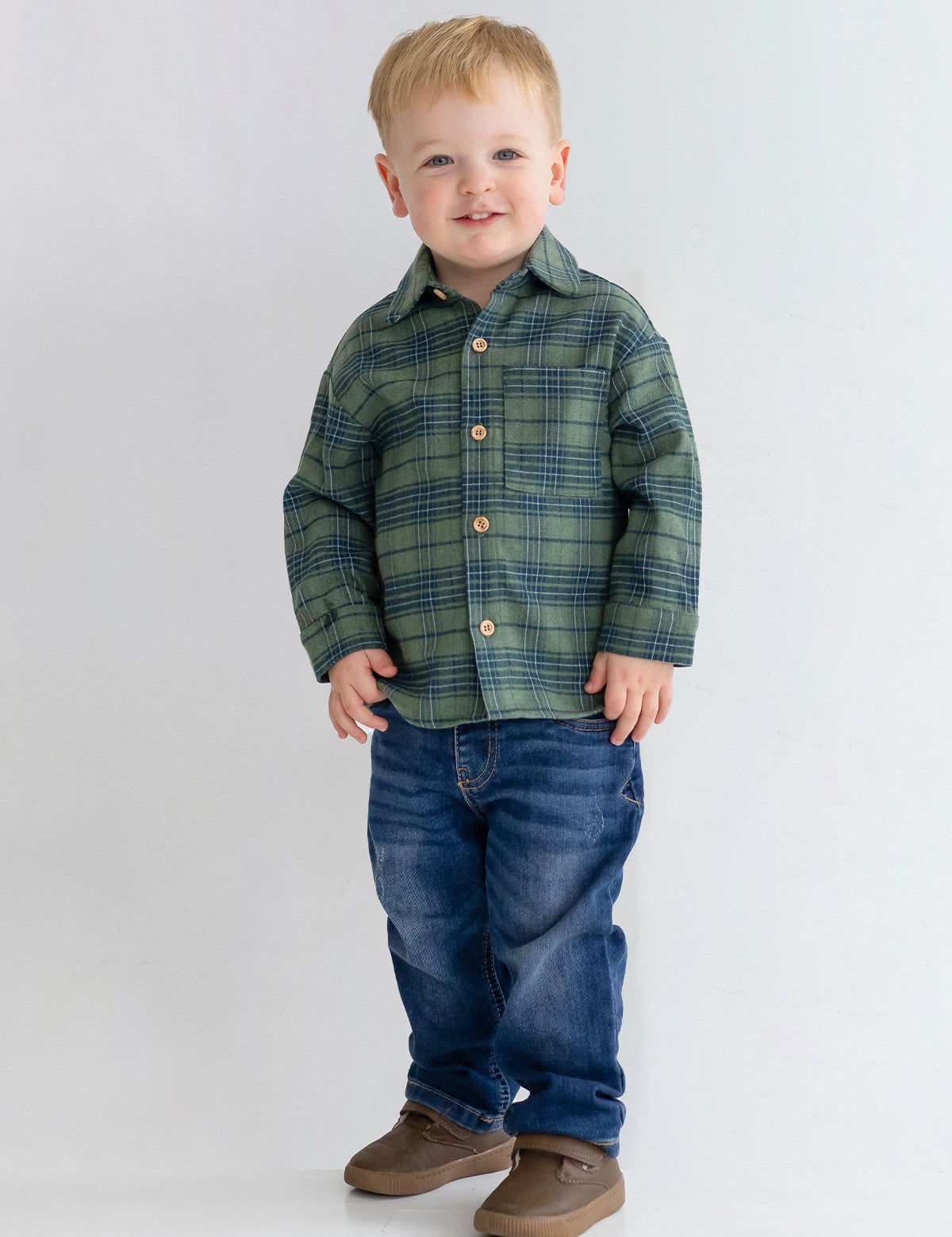 A young child with short blond hair smiles while standing against a plain light background, wearing the Beckett and Bear Cozy Creatures Brothers Top, blue jeans, and brown shoes.