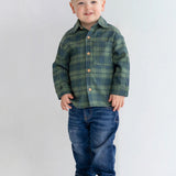A young child with short blond hair smiles while standing against a plain light background, wearing the Beckett and Bear Cozy Creatures Brothers Top, blue jeans, and brown shoes.