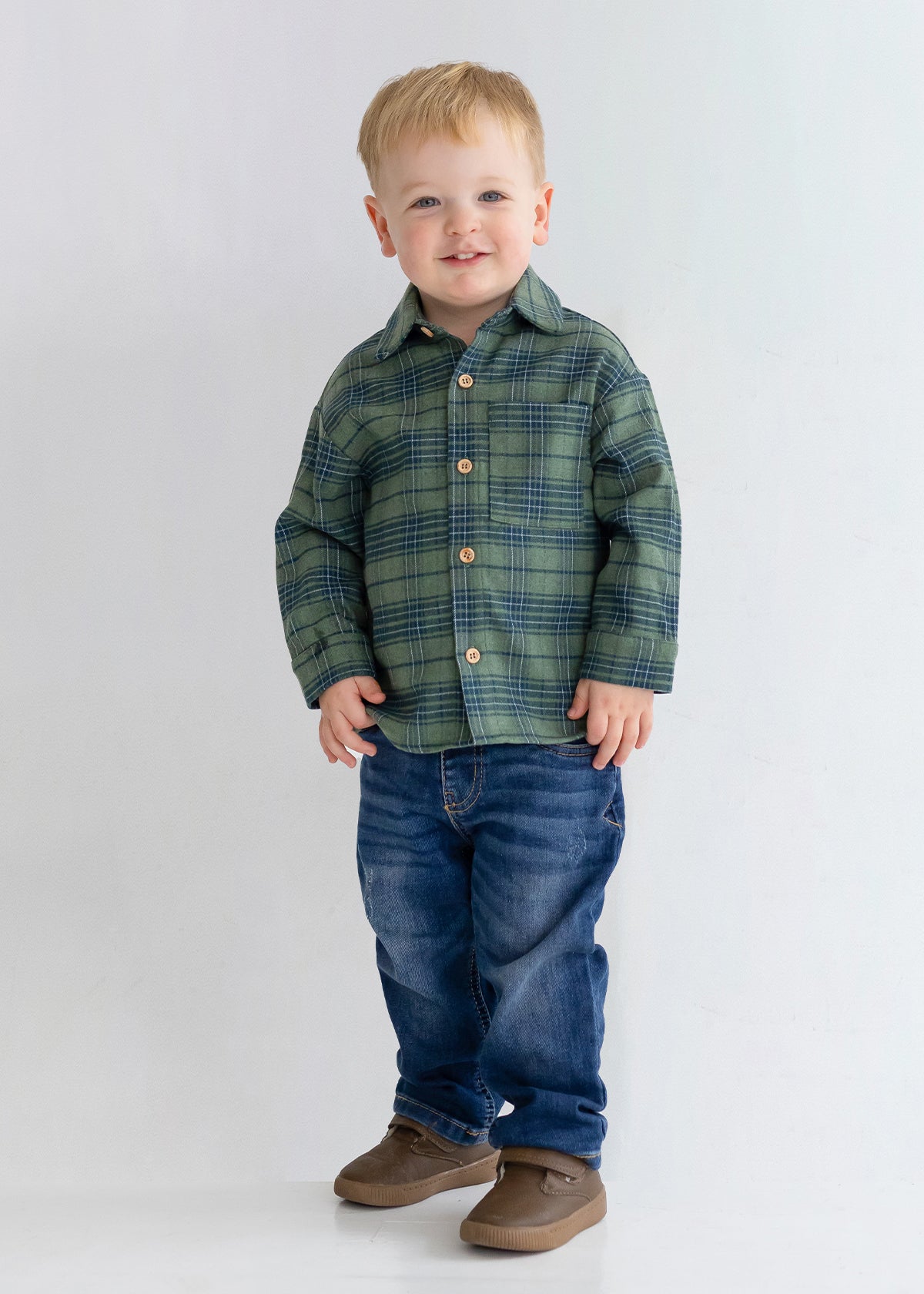 A young child with short blond hair smiles while standing against a plain light background, wearing the Beckett and Bear Cozy Creatures Brothers Top, blue jeans, and brown shoes.