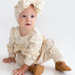 A baby wearing the Mabel and Honey "Breakfast in Bed" cream floral romper with ruffled sleeves, a matching large bow headband, and brown shoes sits on a white floor, gazing slightly to the side.