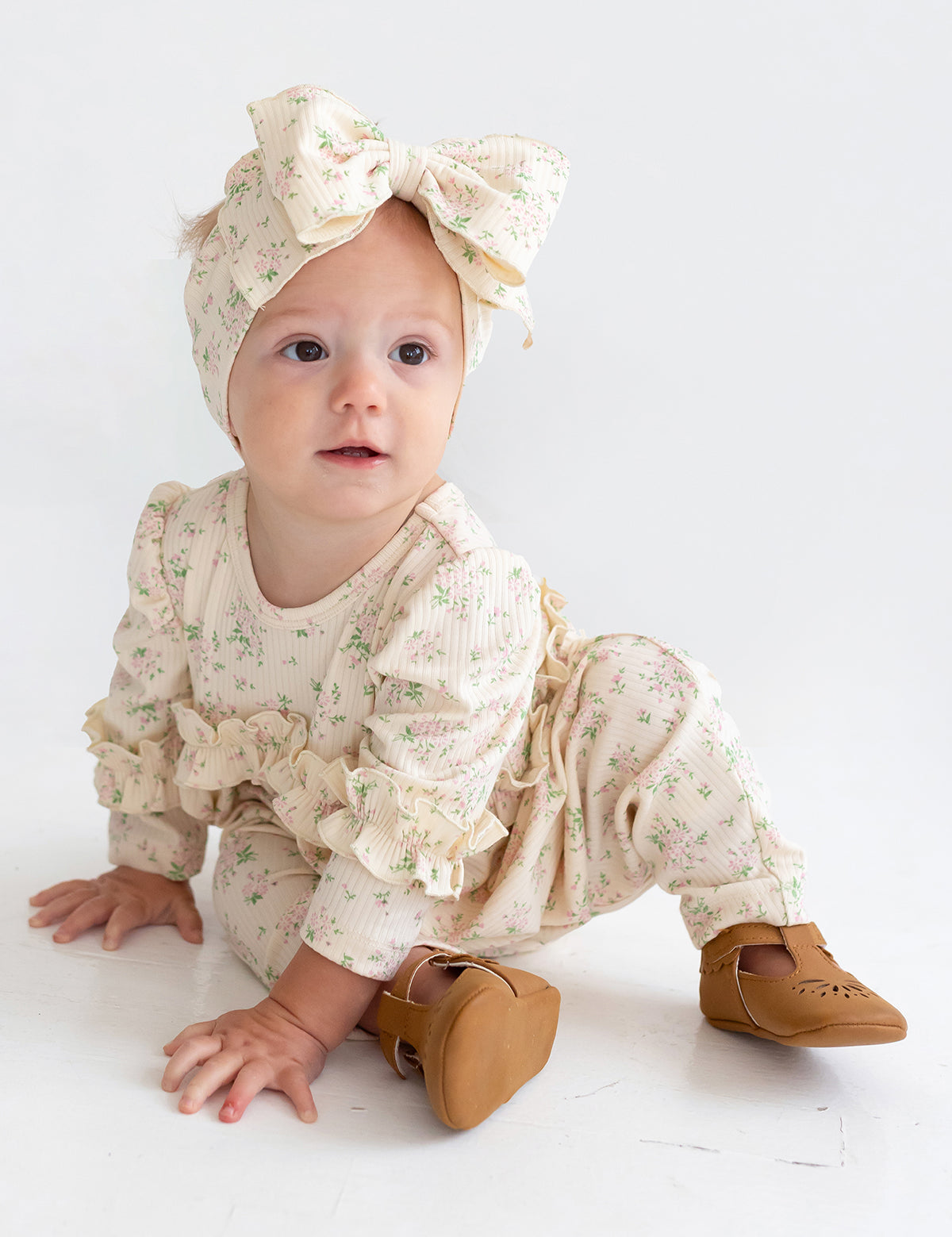 A baby wearing the Mabel and Honey "Breakfast in Bed" cream floral romper with ruffled sleeves, a matching large bow headband, and brown shoes sits on a white floor, gazing slightly to the side.