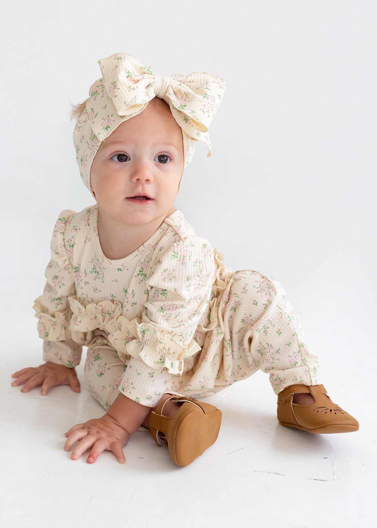 A baby wearing the Mabel and Honey "Breakfast in Bed" cream floral romper with ruffled sleeves, a matching large bow headband, and brown shoes sits on a white floor, gazing slightly to the side.