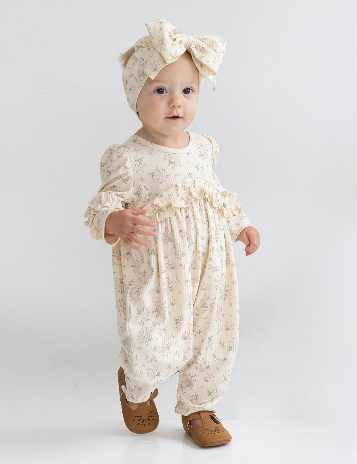 A toddler wears the Mabel and Honey "Breakfast in Bed Romper," a cream floral outfit with ruffled sleeves, paired with a matching large bow headband and brown shoes, standing against a plain white background.