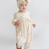 A toddler wears the Mabel and Honey "Breakfast in Bed Romper," a cream floral outfit with ruffled sleeves, paired with a matching large bow headband and brown shoes, standing against a plain white background.