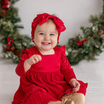 A smiling baby girl wearing the Isobella and Chloe Merry 'n Bright Red Velvet Dress and matching headband sits by a festive wreath. She pairs the outfit with gold shoes and reaches forward with one hand.