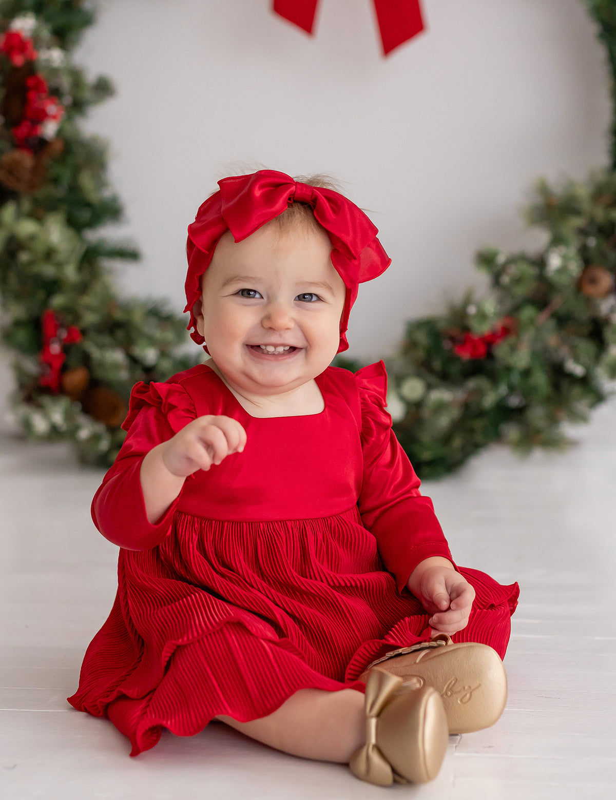 A smiling baby girl wearing the Isobella and Chloe Merry 'n Bright Red Velvet Dress and matching headband sits by a festive wreath. She pairs the outfit with gold shoes and reaches forward with one hand.