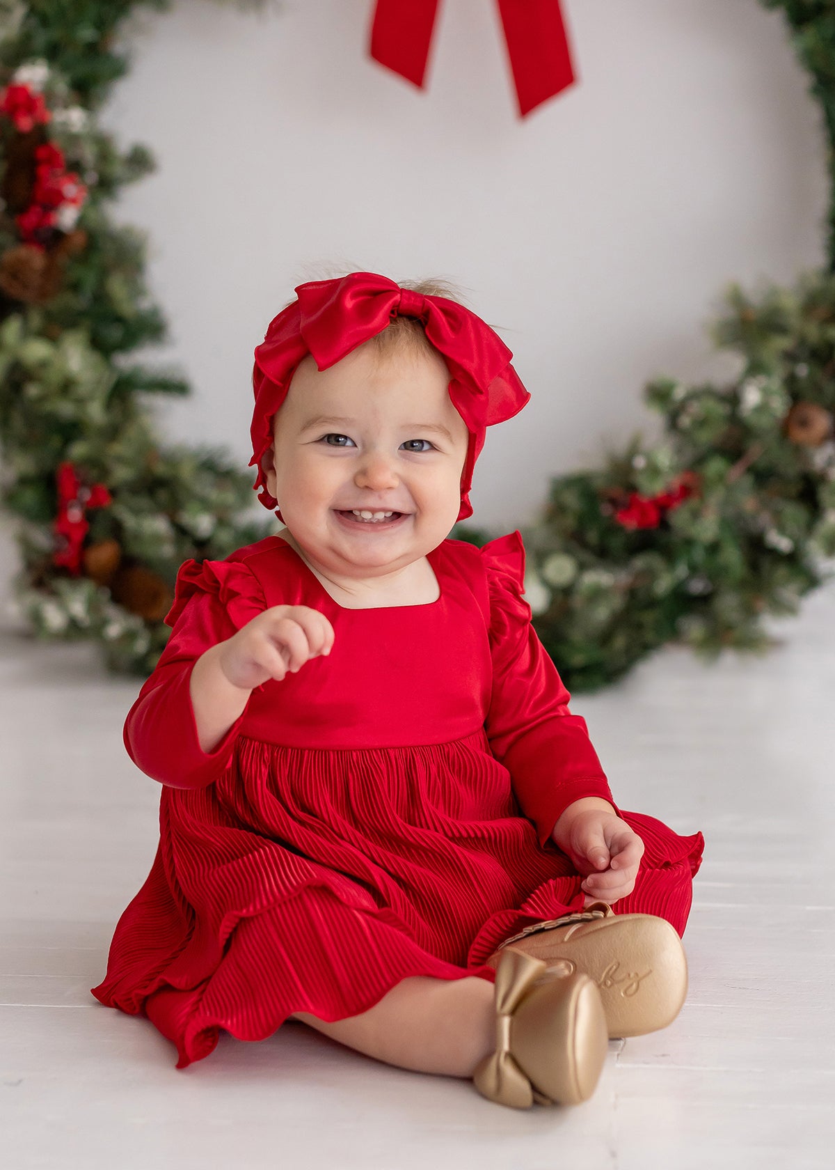 A smiling baby girl wearing the Isobella and Chloe Merry 'n Bright Red Velvet Dress and matching headband sits by a festive wreath. She pairs the outfit with gold shoes and reaches forward with one hand.