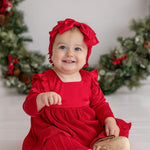 A smiling baby in the Merry 'n Bright Red Velvet Dress by Isobella and Chloe and a matching headband sits before a festive holiday wreath, wearing gold shoes labeled "Baby." Greenery on the white background creates a cheerful holiday scene.