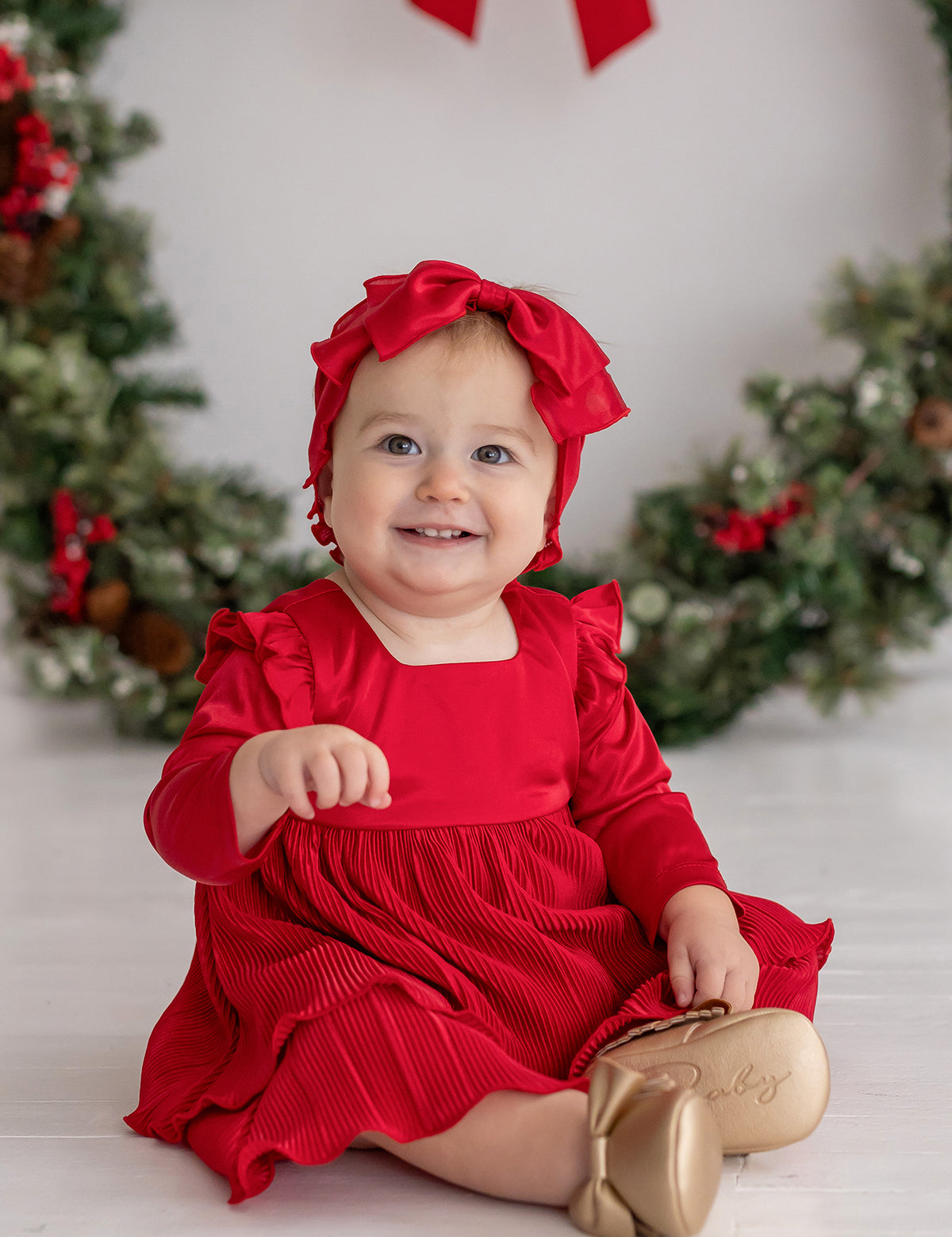A smiling baby in the Merry 'n Bright Red Velvet Dress by Isobella and Chloe and a matching headband sits before a festive holiday wreath, wearing gold shoes labeled "Baby." Greenery on the white background creates a cheerful holiday scene.