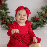 A smiling baby in the Merry 'n Bright Red Velvet Dress by Isobella and Chloe and a matching headband sits before a festive holiday wreath, wearing gold shoes labeled "Baby." Greenery on the white background creates a cheerful holiday scene.