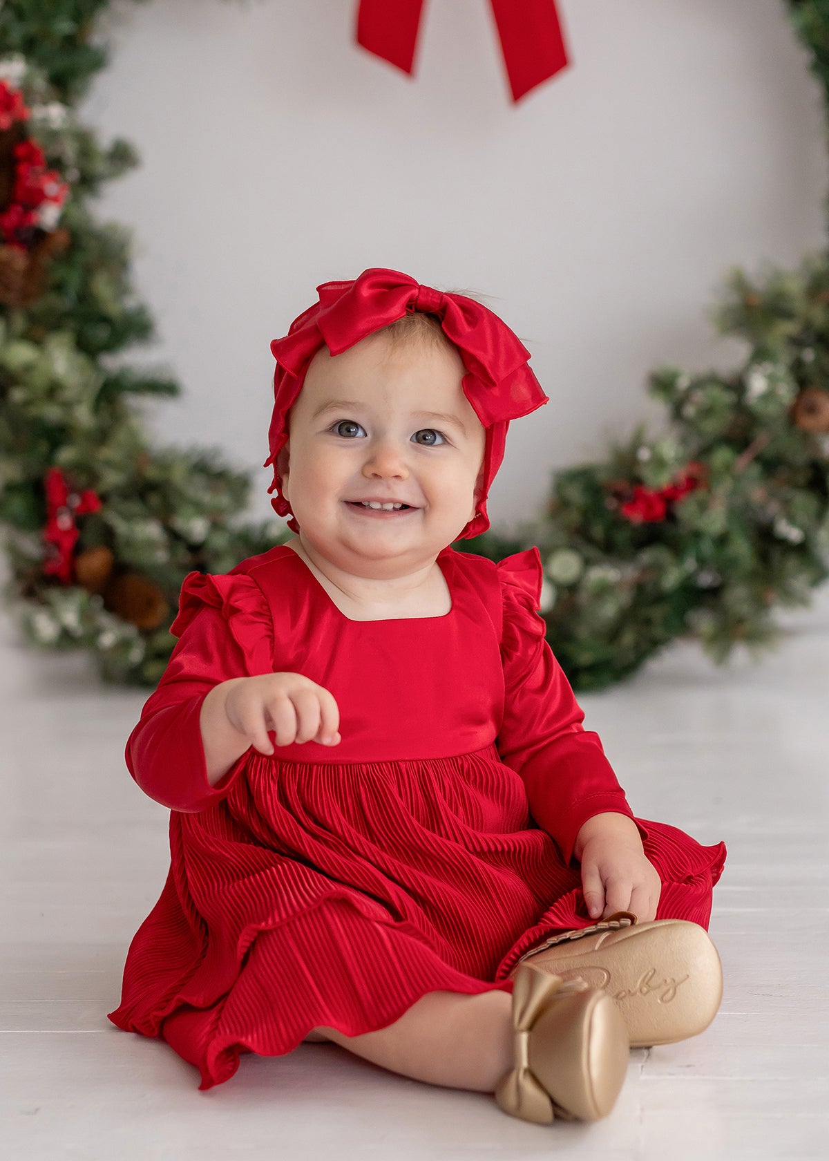 A smiling baby in the Merry 'n Bright Red Velvet Dress by Isobella and Chloe and a matching headband sits before a festive holiday wreath, wearing gold shoes labeled "Baby." Greenery on the white background creates a cheerful holiday scene.