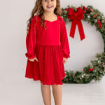 A young girl with long curly hair smiles indoors, wearing the Isobella and Chloe Merry n Bright Velvet Dress—red with long sleeves, natural waist, and pleated skirt—plus beige shoes, standing in front of a large wreath with a red bow.
