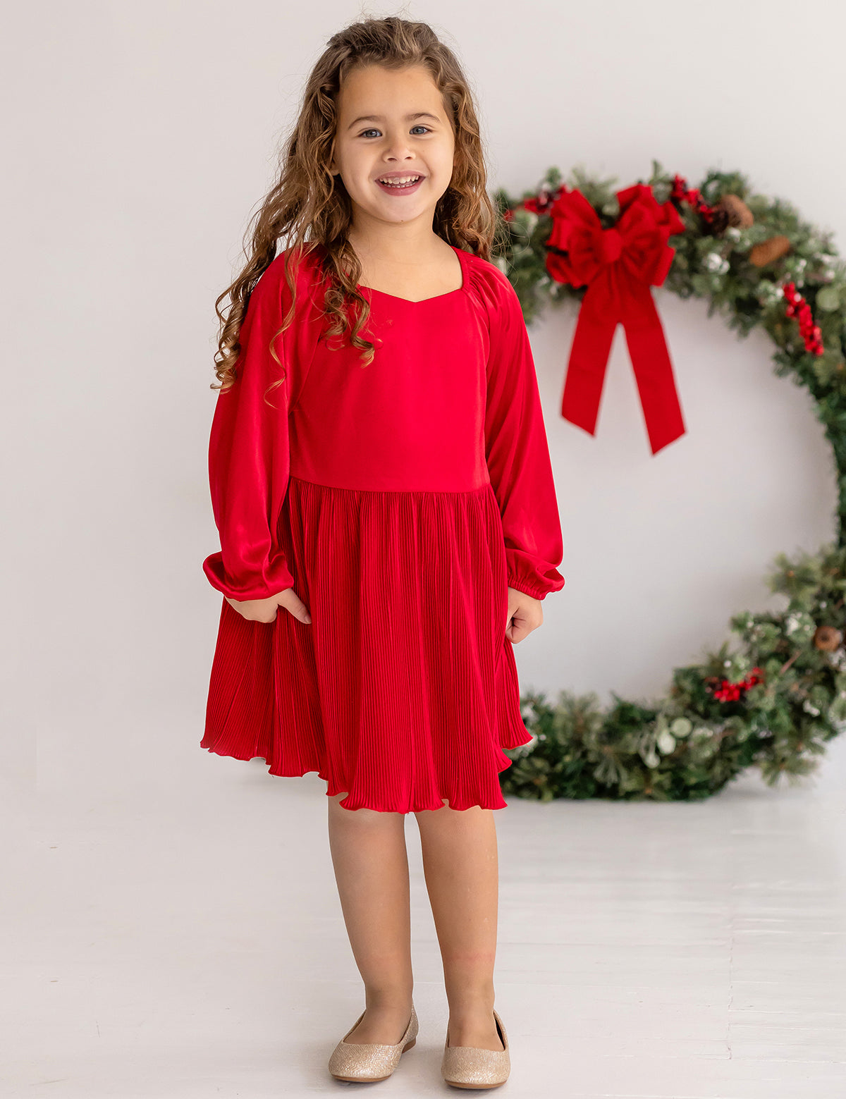 A young girl with long curly hair smiles indoors, wearing the Isobella and Chloe Merry n Bright Velvet Dress—red with long sleeves, natural waist, and pleated skirt—plus beige shoes, standing in front of a large wreath with a red bow.
