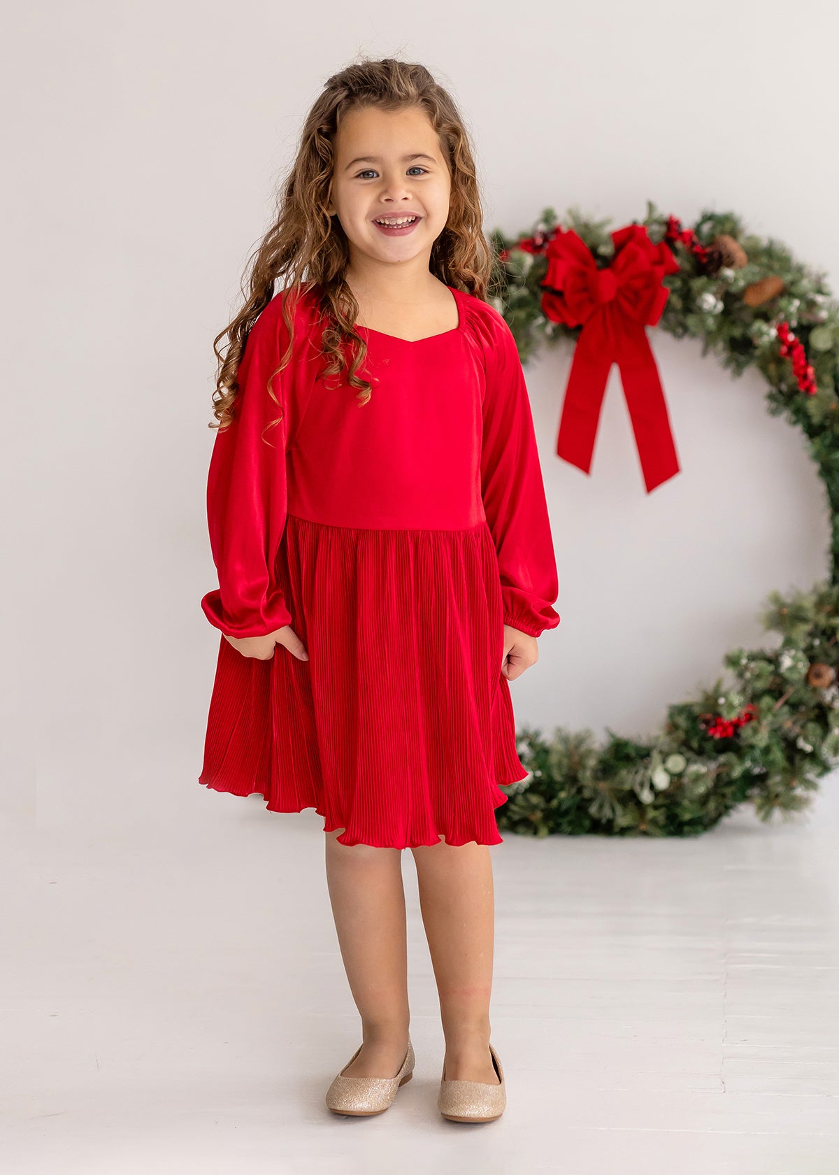 A young girl with long curly hair smiles indoors, wearing the Isobella and Chloe Merry n Bright Velvet Dress—red with long sleeves, natural waist, and pleated skirt—plus beige shoes, standing in front of a large wreath with a red bow.