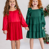 Two young girls stand hand in hand, smiling. One wears the Isobella and Chloe Merry n Bright Velvet Dress in red; the other, a green ruffled dress. Both have long hair and light-colored shoes—perfect for festive holiday gatherings.