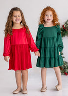 Two young girls stand hand in hand, smiling. One wears the Isobella and Chloe Merry n Bright Velvet Dress in red; the other, a green ruffled dress. Both have long hair and light-colored shoes—perfect for festive holiday gatherings.