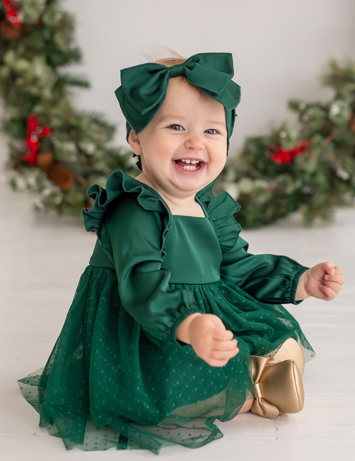 A smiling baby wears the Isobella and Chloe Mistletoe Emerald Dress with a matching headband and gold shoes, surrounded by holiday wreaths with red ribbons—perfect for festive celebrations.