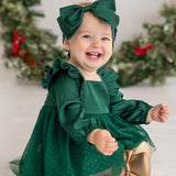 A smiling baby wears the Isobella and Chloe Mistletoe Emerald Dress with a matching headband and gold shoes, surrounded by holiday wreaths with red ribbons—perfect for festive celebrations.