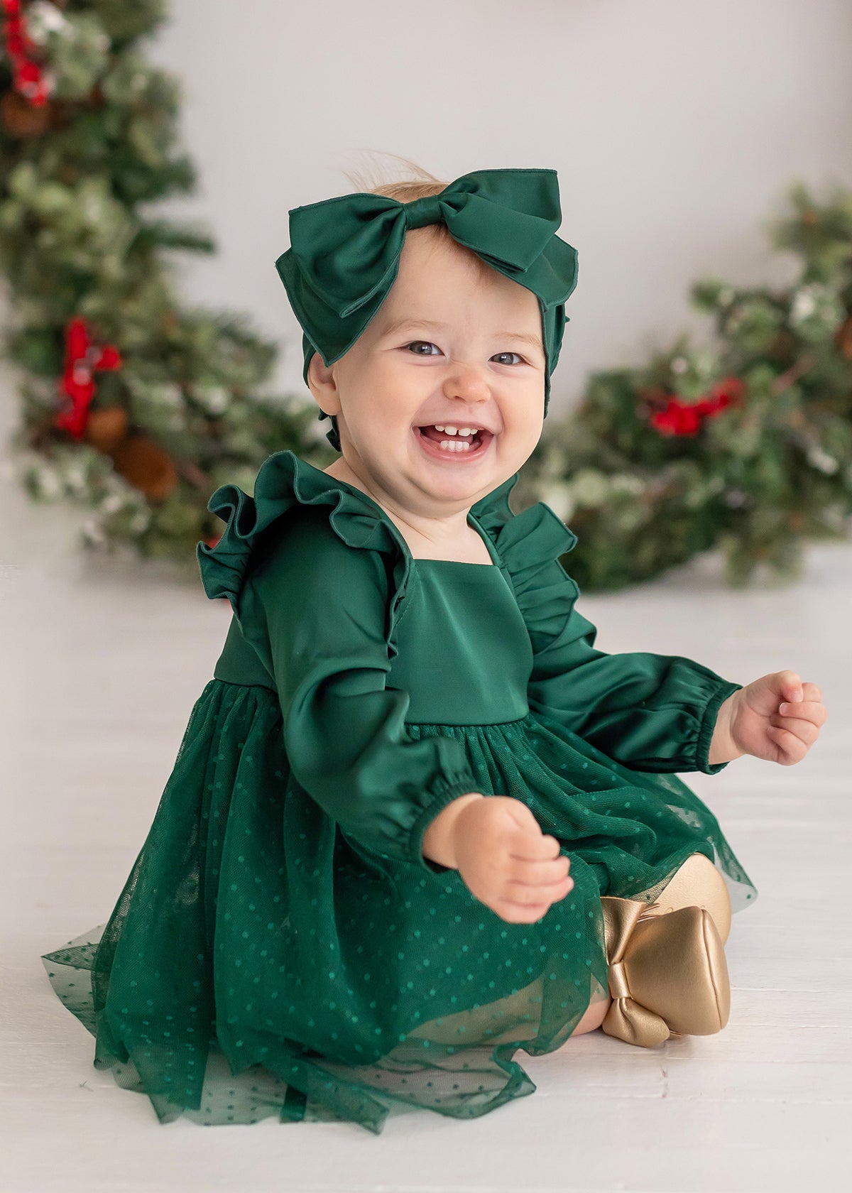 A smiling baby wears the Isobella and Chloe Mistletoe Emerald Dress with a matching headband and gold shoes, surrounded by holiday wreaths with red ribbons—perfect for festive celebrations.