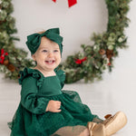 A smiling baby girl wears the Isobella and Chloe Mistletoe Emerald Dress with a matching bow, gold shoes, and sheer polka-dot stockings as she sits before a festive wreath—perfect attire for holiday celebrations.