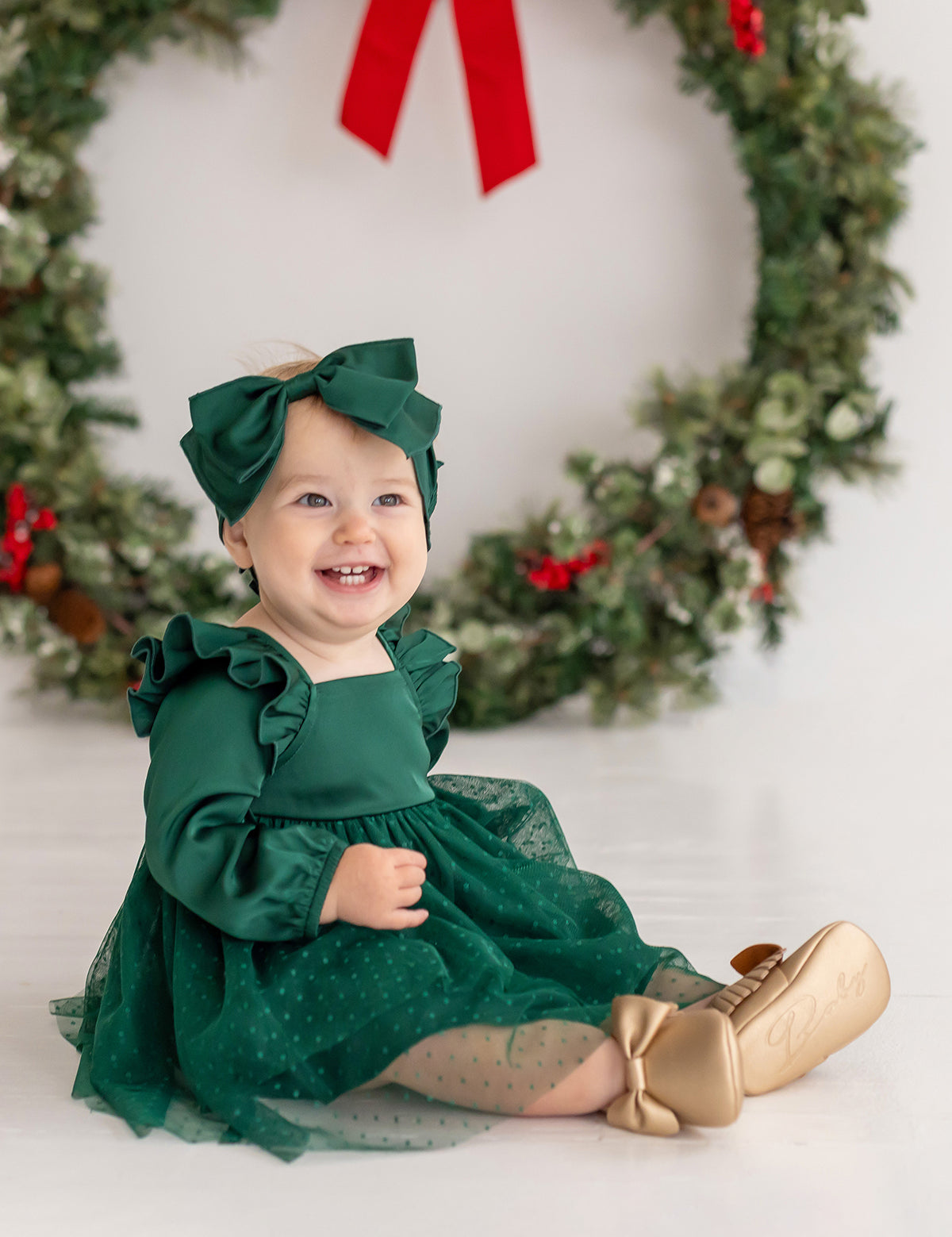 A smiling baby girl wears the Isobella and Chloe Mistletoe Emerald Dress with a matching bow, gold shoes, and sheer polka-dot stockings as she sits before a festive wreath—perfect attire for holiday celebrations.