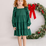 A young girl with curly red hair wears the Isobella and Chloe Mistletoe Emerald Green Tiered Dress and beige shoes, standing indoors on a white floor. A festive wreath with a red bow hangs in the background.