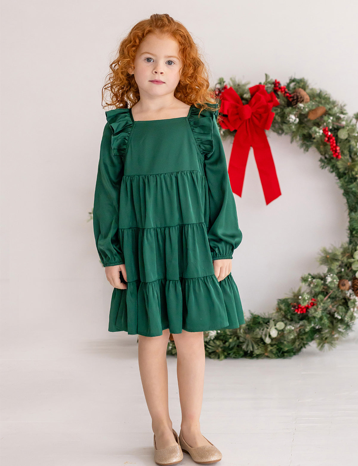 A young girl with curly red hair wears the Isobella and Chloe Mistletoe Emerald Green Tiered Dress and beige shoes, standing indoors on a white floor. A festive wreath with a red bow hangs in the background.