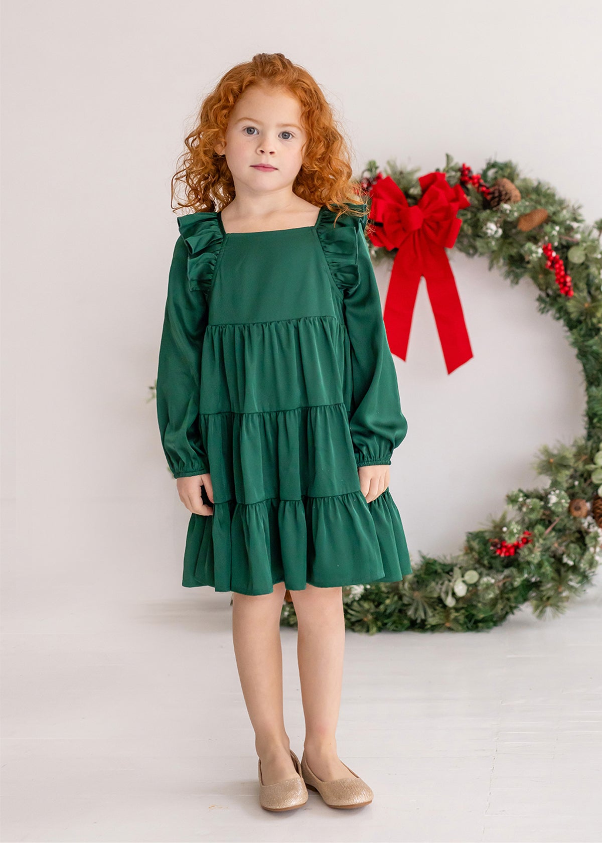 A young girl with curly red hair wears the Isobella and Chloe Mistletoe Emerald Green Tiered Dress and beige shoes, standing indoors on a white floor. A festive wreath with a red bow hangs in the background.