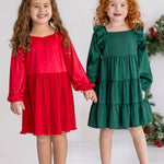 Two young girls stand side by side, smiling and holding hands. One wears a red holiday party dress, while the other wears the Isobella and Chloe Mistletoe Emerald Green Tiered Dress. Both have curly hair, beige shoes, and a festive wreath in the background.