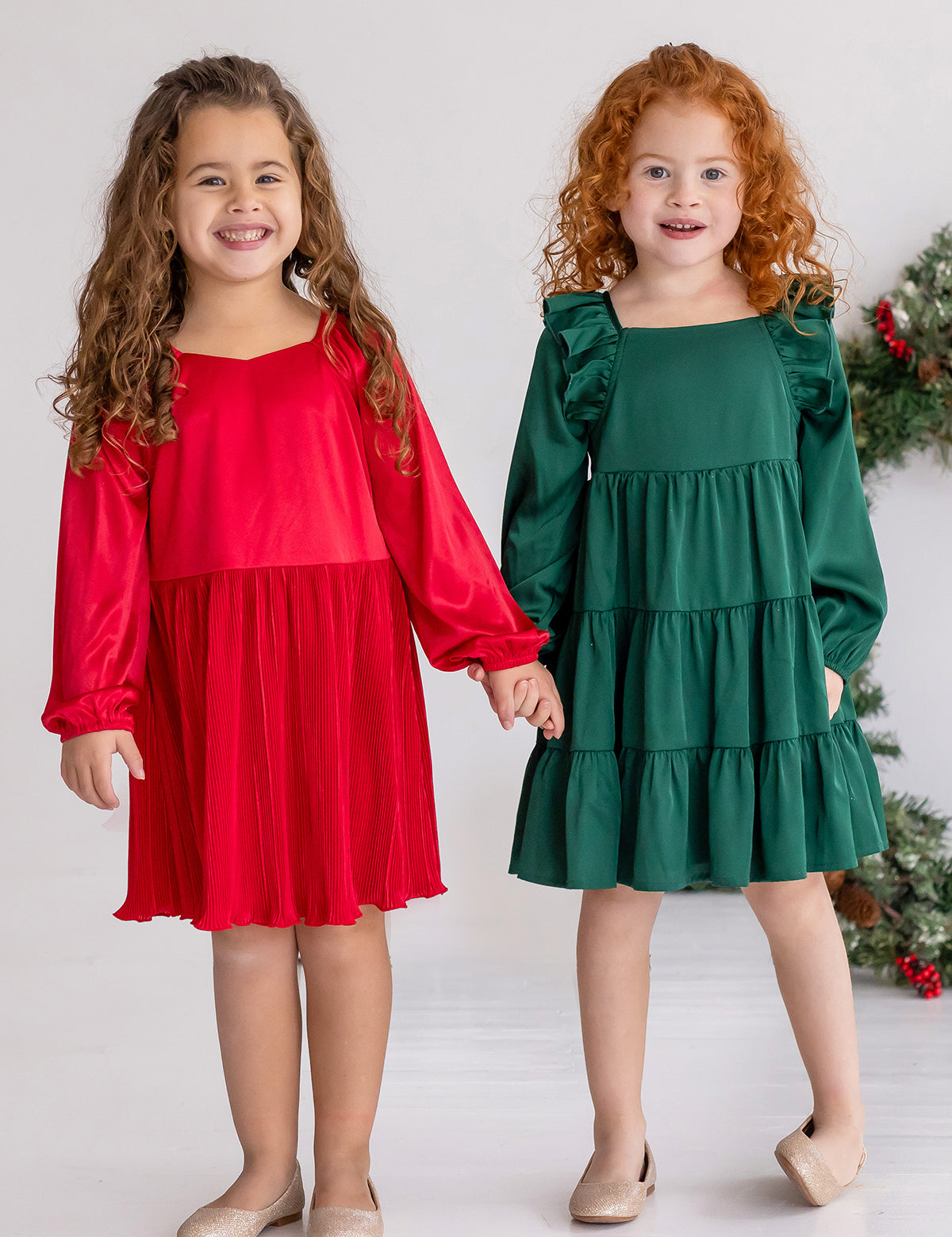 Two young girls stand side by side, smiling and holding hands. One wears a red holiday party dress, while the other wears the Isobella and Chloe Mistletoe Emerald Green Tiered Dress. Both have curly hair, beige shoes, and a festive wreath in the background.