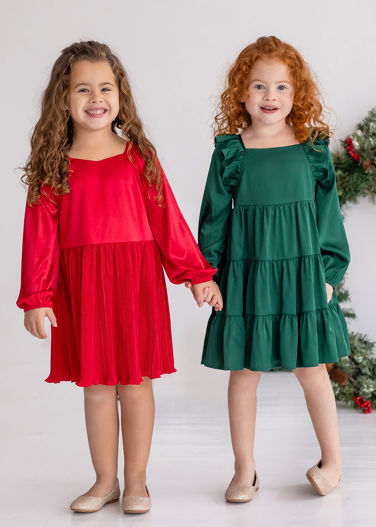 Two young girls stand side by side, smiling and holding hands. One wears a red holiday party dress, while the other wears the Isobella and Chloe Mistletoe Emerald Green Tiered Dress. Both have curly hair, beige shoes, and a festive wreath in the background.