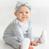 A baby wears the Mabel and Honey "Hannah Romper," featuring a blue and white floral print with ruffled shoulders, paired with a matching bow headband and white shoes while sitting on a white floor, smiling up.