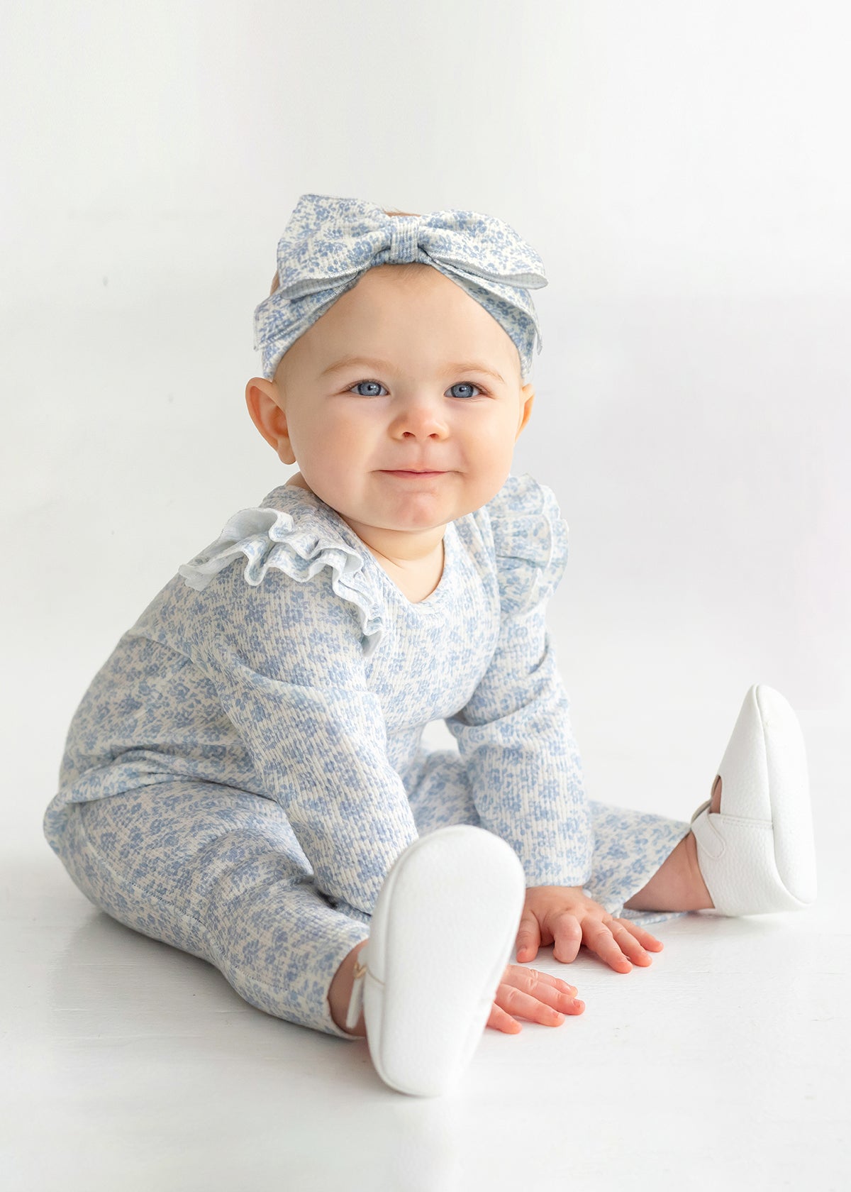 A baby wears the Mabel and Honey "Hannah Romper," featuring a blue and white floral print with ruffled shoulders, paired with a matching bow headband and white shoes while sitting on a white floor, smiling up.