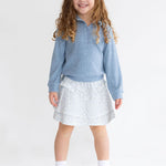 A young girl with long curly hair smiles against a plain white backdrop, wearing Mabel and Honey's Hannah Two Piece Set: a light blue long-sleeve pullover and floral skirt, paired with white socks and sneakers.