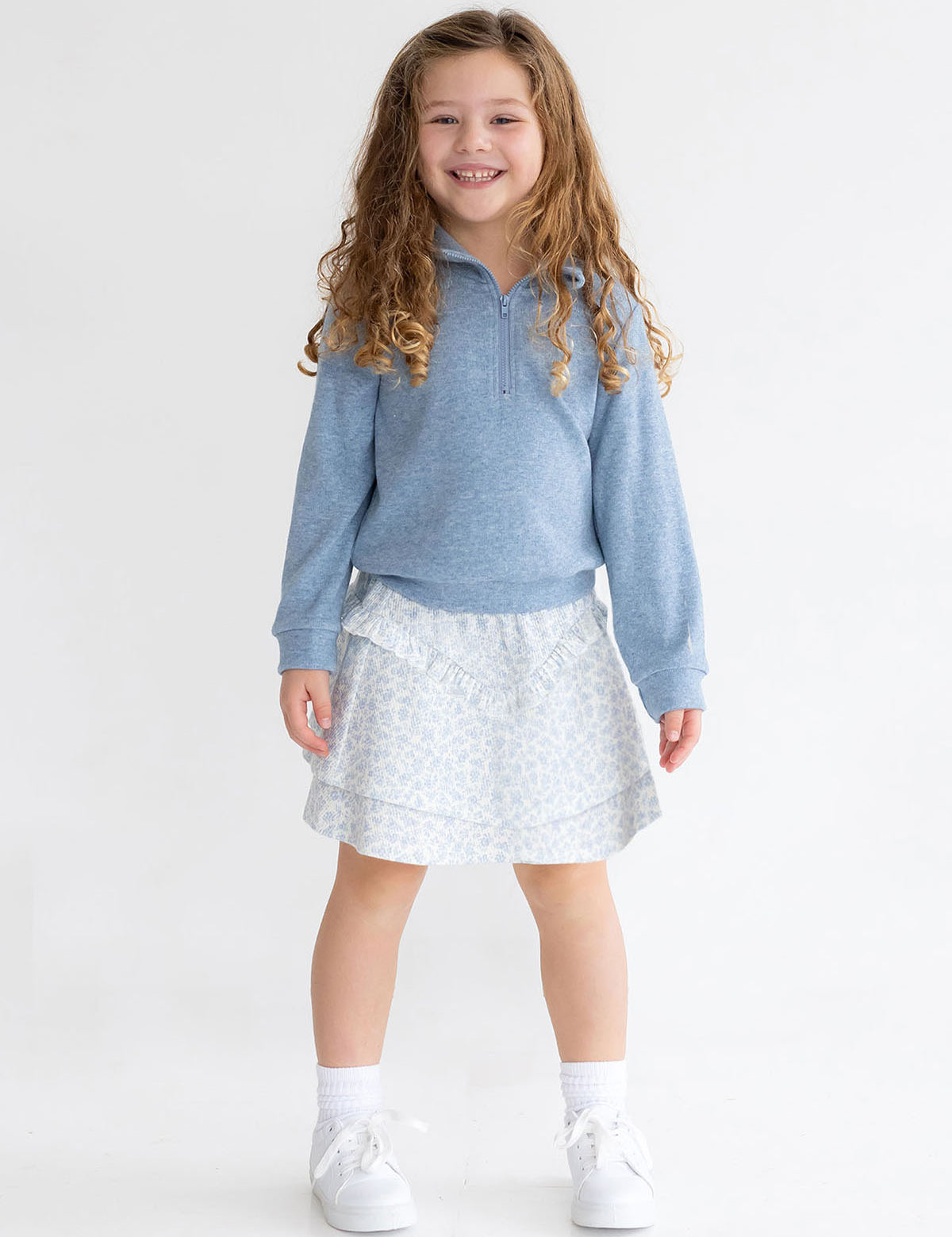 A young girl with long curly hair smiles against a plain white backdrop, wearing Mabel and Honey's Hannah Two Piece Set: a light blue long-sleeve pullover and floral skirt, paired with white socks and sneakers.