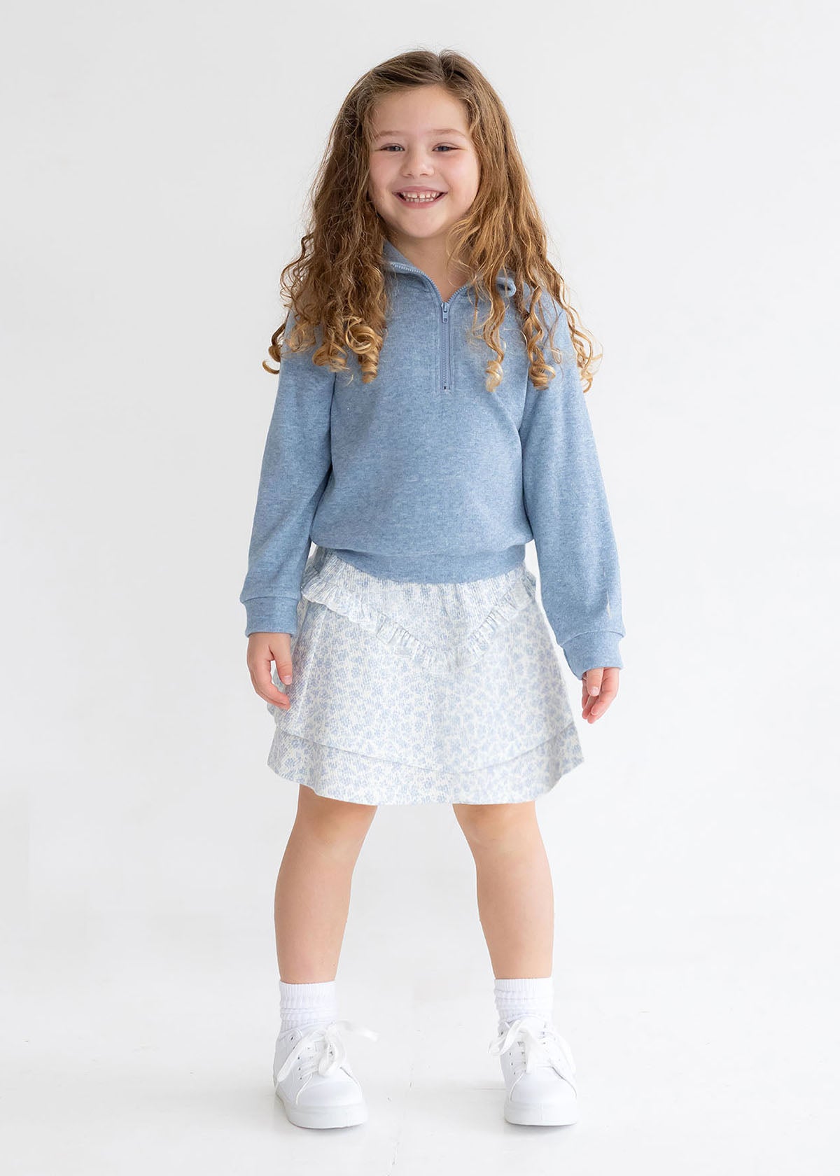 A young girl with long curly hair smiles against a plain white backdrop, wearing Mabel and Honey's Hannah Two Piece Set: a light blue long-sleeve pullover and floral skirt, paired with white socks and sneakers.