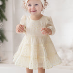 A young toddler smiles indoors wearing the Isobella and Chloe Heart of Gold Little Big Girl Dress—an ivory tulle dress with a layered ruffle skirt—paired with bow shoes and a headband, surrounded by greenery and white decor.