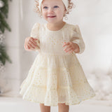 A young toddler smiles indoors wearing the Isobella and Chloe Heart of Gold Little Big Girl Dress—an ivory tulle dress with a layered ruffle skirt—paired with bow shoes and a headband, surrounded by greenery and white decor.